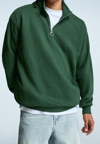 Sudadera polar de color verde oscuro con cuello simulado, cierre de cremallera y puños acanalados. Lleva puesta sobre una camiseta blanca, combinada con vaqueros azul claro.