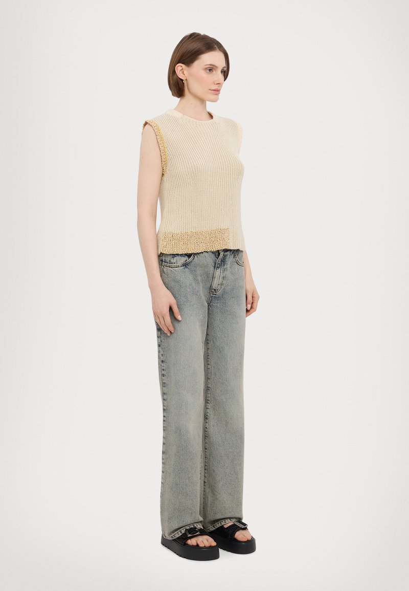 Beige gebreid mouwloos topje met een tekstuur aan de onderrand, gecombineerd met lichtblauwe high-waisted wijde jeans en zwarte platform sandalen.