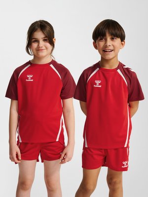 Dos niños sonrientes con camisetas deportivas rojas a juego y pantalones cortos con detalles blancos, de pie frente a un fondo blanco liso.