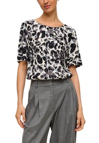 Kurzärmlige Bluse mit einem Leopardenmuster in Schwarz, Grau und Weiß; leichter Stoff mit gefälteltem Detail an der Vorderseite. Kombiniert mit grauen, maßgeschneiderten Hosen.
