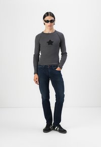 Suéter gris de manga larga con un parche de estrella negra, combinado con jeans azul oscuro y zapatillas negras con rayas blancas. Lleva puestas gafas de sol claras.