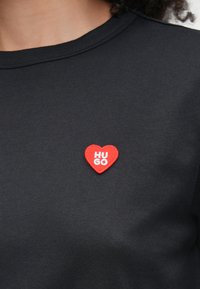 Melnas kokvilnas T-krekls ar sarkanu sirdi formas ieliktni un uzrakstu "HUGO". Krekls ir ar apaļu kakla izgriezumu un gludu tekstūru.