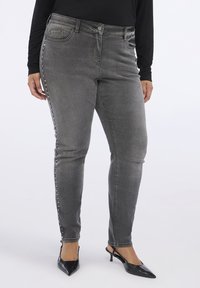 Jeans ajustados grises de cintura alta, con un ligero desgaste, detalles de botones en los laterales y una textura de denim elástico. Combinados con tacones negros puntiagudos.