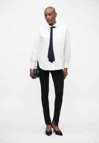 Witte button-up blouse met een zak, gecombineerd met zwarte slim-fit broek en zwarte hakschoenen. Model houdt een kleine zwarte handtas vast.