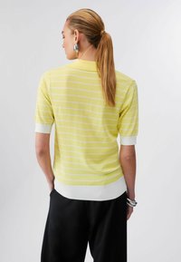 Femme avec queue de cheval portant un t-shirt à manches courtes rayé jaune et blanc, pantalon noir, boucles d'oreilles argentées et bracelets, tournée de dos à la caméra.