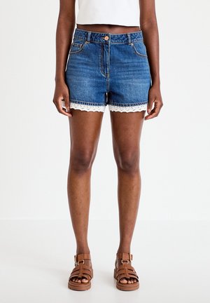 Woman wearing high-waisted blue denim shorts with white lace trim, paired with brown strappy sandals and a white cropped top.