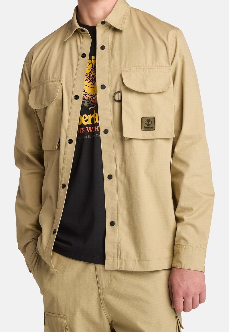 Homme portant une veste-chemise beige boutonnée avec deux poches poitrine à rabat par-dessus un t-shirt noir graphique et un pantalon beige assorti.