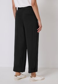 Pantalons larges noirs avec une taille élastique, deux poches arrière, et un tissu lisse et léger, associés à des baskets blanches.