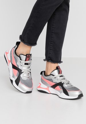 Osoba nosząca srebrne, różowe, czarne i szare siatkowe buty Puma z białymi podeszwami oraz czarne skrócone jeansy na jednolitym tle.