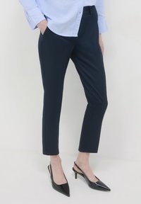 Frau in navyblauen, maßgeschneiderten Knöchelhosen, lichtblauem Hemd und schwarzen Slingback-Highheels mit spitzem Zehenteil, stehend vor einem weißen Hintergrund.