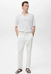 Polo shirt bianco a maniche corte in maglia con colletto a coste e motivo geometrico, abbinato a pantaloni bianchi su misura e sandali neri.