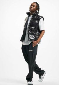 Black shiny puffer vest with a high collar over a white t-shirt, paired with loose black pants featuring a logo, and gray and black sneakers.