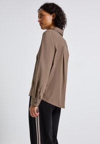 Donna con capelli ricci che indossa una maglietta marrone a maniche lunghe e pantaloni neri con strisce bianche, rivolta a sinistra su uno sfondo grigio chiaro.