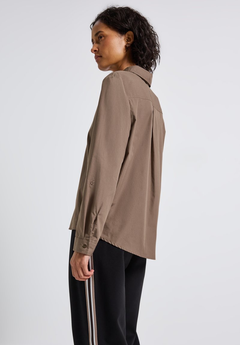 Donna con capelli ricci che indossa una maglietta marrone a maniche lunghe e pantaloni neri con strisce bianche, rivolta a sinistra su uno sfondo grigio chiaro.