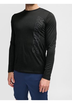 Homme portant un t-shirt noir à manches longues avec le texte "HUMMEL" sur le côté et un short bleu, debout devant un fond uni.