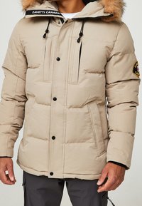 Chaqueta de plumón beige con capucha forrada de piel, herrajes negros, dos bolsillos frontales y un parche con logo en la manga. Tejido texturizado con un diseño acolchado.