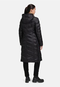 Zwarte pufferjas met een chevron-quiltontwerp, capuchon en zijritsen. Gladde textuur; middenlengte, gecombineerd met zwarte broeken en stevige laarzen.