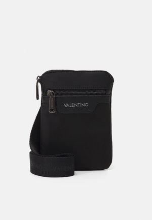 Zwarte Valentino crossbodytas met een voorvak met rits, zilverkleurige ritstrekkers en een verstelbare, gebrande riem tegen een lichte achtergrond.