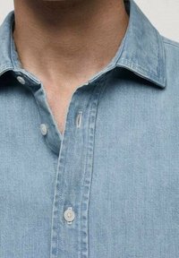 Chemise en denim bleu clair avec col classique, patte de boutonnage et coutures visibles. Texture douce avec un léger effet délavé.