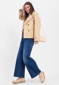 Beige Doppelreiher-Trenchcoat, weite blaue Jeans und beigefarbene Slipper mit dicker Sohle; eine goldene Kettenhandtasche hängt von der Schulter.