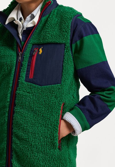 Πράσινο φλις γιλέκο με υφή, ναυτικές λεπτομέρειες και τσέπη, κόκκινος φερμουάρ. Φορεμένο με μπλε και πράσινη ριγέ μακρυμάνικη μπλούζα.