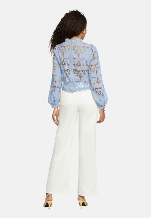 Femme portant un chemisier en dentelle légère bleu clair à manches longues et un pantalon blanc ample taille haute, debout de dos.