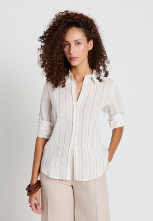 Frau mit lockigem Haar, die ein weiß gestreiftes Hemd mit Knöpfen und beige Hose trägt, steht mit einer Hand in der Tasche und braunen Armbändern am Handgelenk.