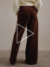 Bruine ribfluwelen broek met wijde pijpen, achterzakken en zichtbare stiksels, gecombineerd met een lichtgekleurde trui met lange mouwen.