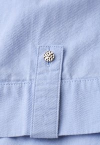 Tissu bleu clair avec texture diagonale et gros plan sur un bouton-pression argenté texturé sur une patte cousue.