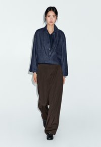 Donna che cammina indossando una giacca cropped blu scuro, top nero, pantaloni marroni larghi, scarpe nere e una lunga collana su uno sfondo chiaro.