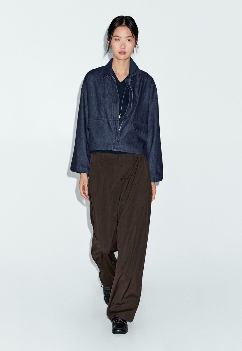 Donna che cammina indossando una giacca cropped blu scuro, top nero, pantaloni marroni larghi, scarpe nere e una lunga collana su uno sfondo chiaro.