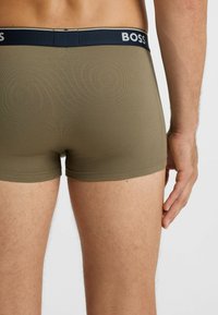 Male wearing olive green fitted boxer briefs with a black waistband labeled "BOSS" on a plain background, hand relaxed at side.