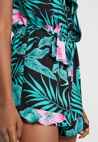 Zwarte playsuit met groen loofpatroon en roze bloemensubstanties. Heeft een gestrikt evenement en een golvende zoom. Gemaakt van een lichte stof.