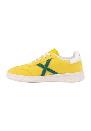 Zapatilla baja amarilla con suela y cordones blancos, con diseño verde en forma de "X" en el lateral y cuello de talón blanco.