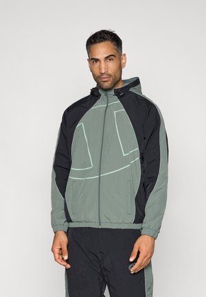Herrenjacke in hellgrün und schwarz, mit einem Reißverschluss, elastischen Bündchen und einer Kapuze. Muster bestehen aus geschwungenen Linien und Kontrastnähten.