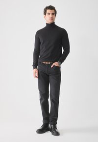Suéter negro de cuello alto, diseño ajustado, combinado con jeans oscuros. El modelo lleva un cinturón marrón y botas negras con cordones, de pie sobre un fondo claro.