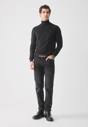 Pulover negru cu guler înalt, design ajustat, asortat cu blugi negri. Modelul poartă o curea maro și bocanci negri cu șireturi, stând pe un fundal deschis.
