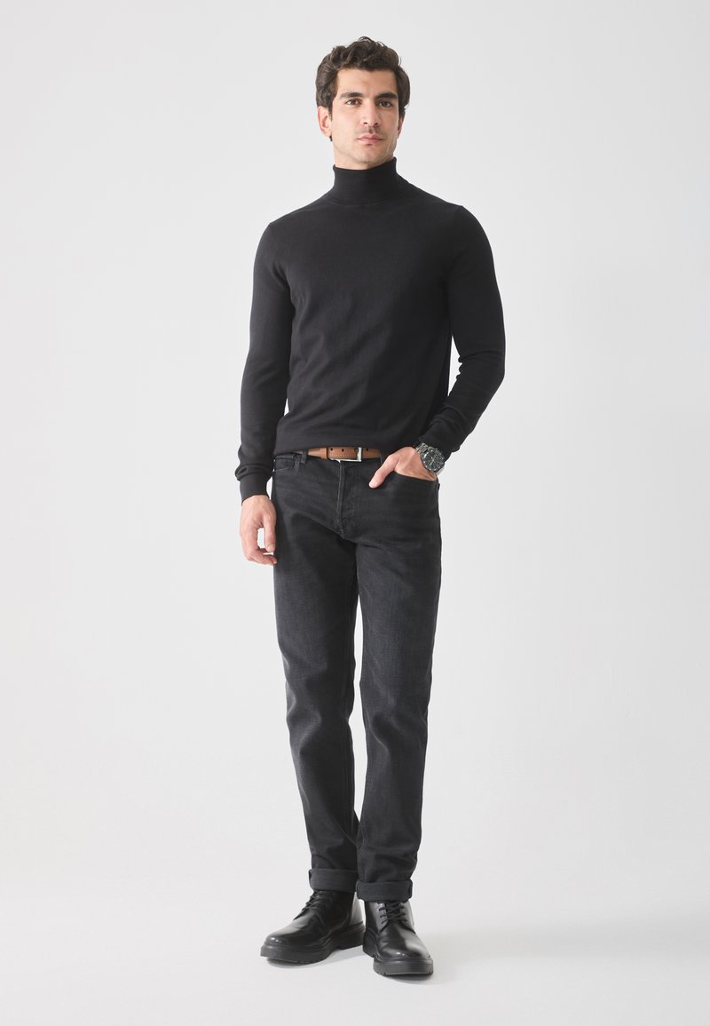 Suéter negro de cuello alto, diseño ajustado, combinado con jeans oscuros. El modelo lleva un cinturón marrón y botas negras con cordones, de pie sobre un fondo claro.