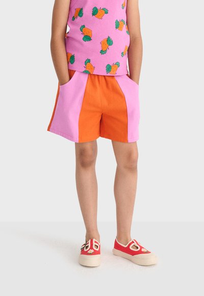 Kind steht mit den Händen in den Taschen einer orange-rosa Shorts, trägt ein ärmelloses rosa Oberteil mit Obstmuster und rote Slip-on-Schuhe mit Blumenmuster.