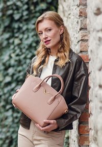 Beige kožená kabelka se dvěma uchy a zlatým akcentem, s dobře strukturovaným tvarem a hladkou texturou. Pozadí tvoří kamenná zeď.