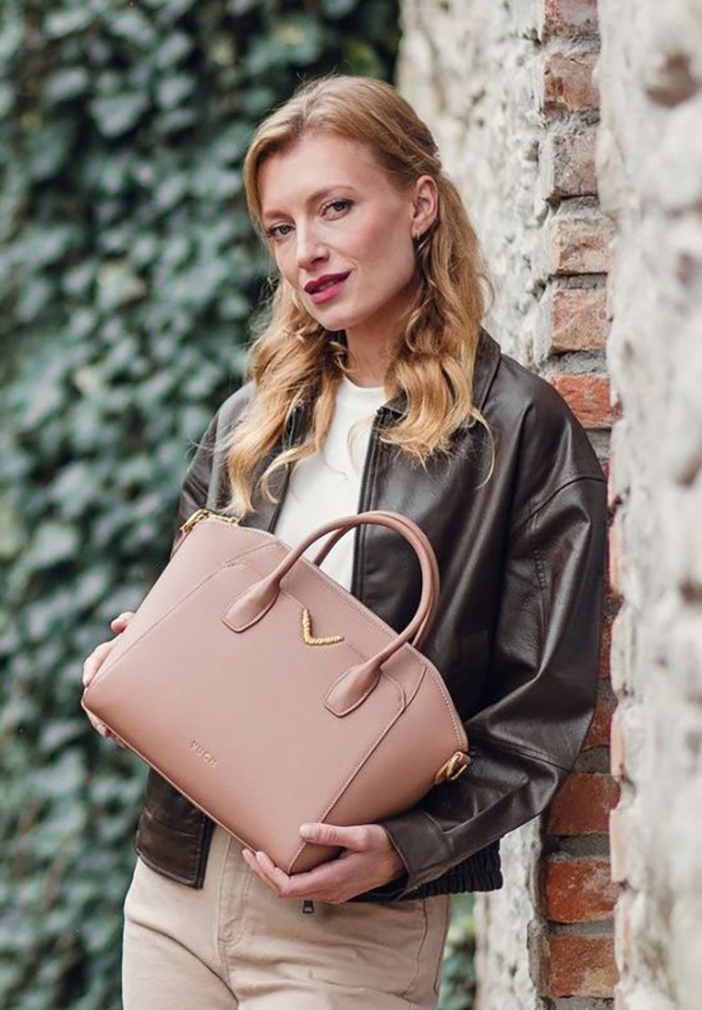 Beige kožená kabelka se dvěma uchy a zlatým akcentem, s dobře strukturovaným tvarem a hladkou texturou. Pozadí tvoří kamenná zeď.