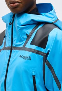 Κοντινό πλάνο ατόμου που φοράει φωτεινό μπλε μπουφάν Columbia Titanium με μαύρες λεπτομέρειες, κουκούλα και εμπρόσθιο φερμουάρ.