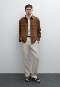 Brun mockapäls knäppjacka med två bröstfickor, vit skjorta under, beige byxor och svarta sandaler med en strukturerad rem.