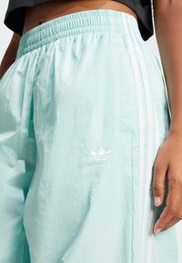 Ľahké športové šortky mätovej farby s elastickým pásom, ktoré majú tri biele pruhy a malé biele logo na nohe.