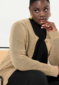 Cardigan beige in maglia aperta con motivo testurizzato, bordi a coste e chiusura con bottoni indossato sopra un top nero. La mano del modello poggia sul mento.