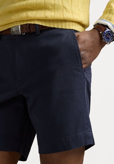 Shorts bleumarin dintr-un material neted, cu buzunare laterale și tiv curat. Asortați cu un pulover galben tricotat și o curea maro.