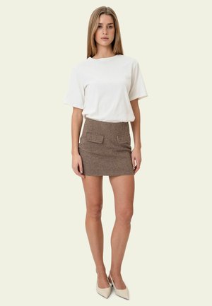 Donna con un top bianco a maniche corte, minigonna a quadri marrone con tasche frontali e tacchi color crema con punta appuntita, in piedi davanti a uno sfondo semplice.