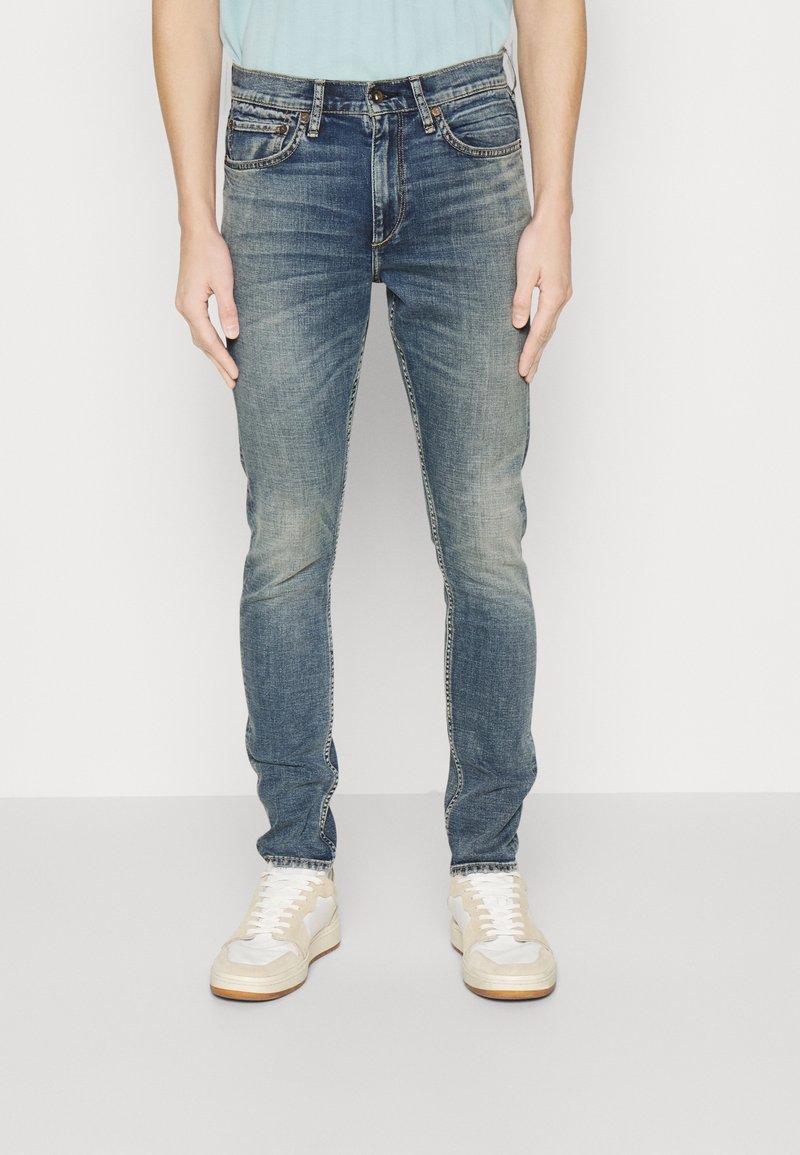 Homme portant un jean slim bleu délavé et des baskets blanches, debout devant un fond clair uni.