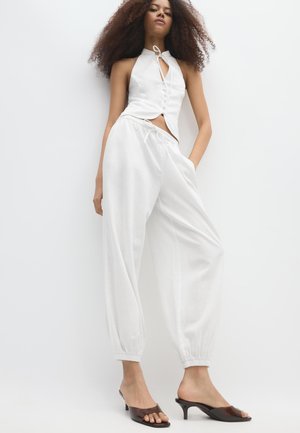 Frau mit lockigem Haar, die ein ärmelloses weißes Neckholder-Top und lockere weiße Hosen trägt und vor einem einfarbigen weißen Hintergrund steht.