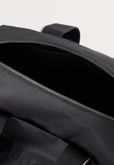 Sac à dos en tissu noir avec une fermeture éclair argentée partiellement ouverte, montrant l'intérieur vide et la poignée supérieure sur un fond clair.